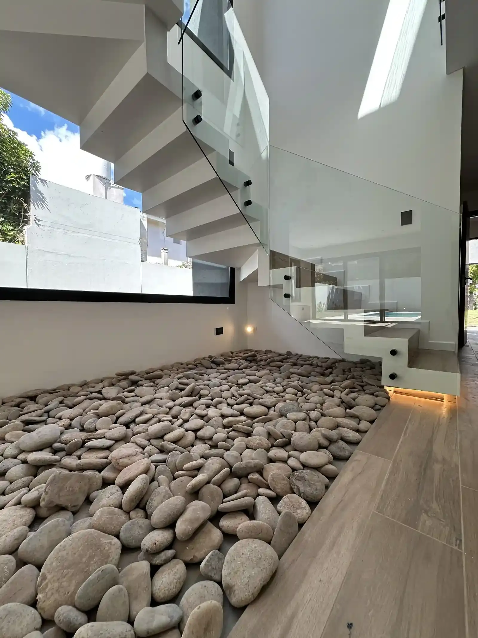 Escalera de madera y vidrio de Casa Adrogué sobre cantero interior de canto rodado, con doble altura