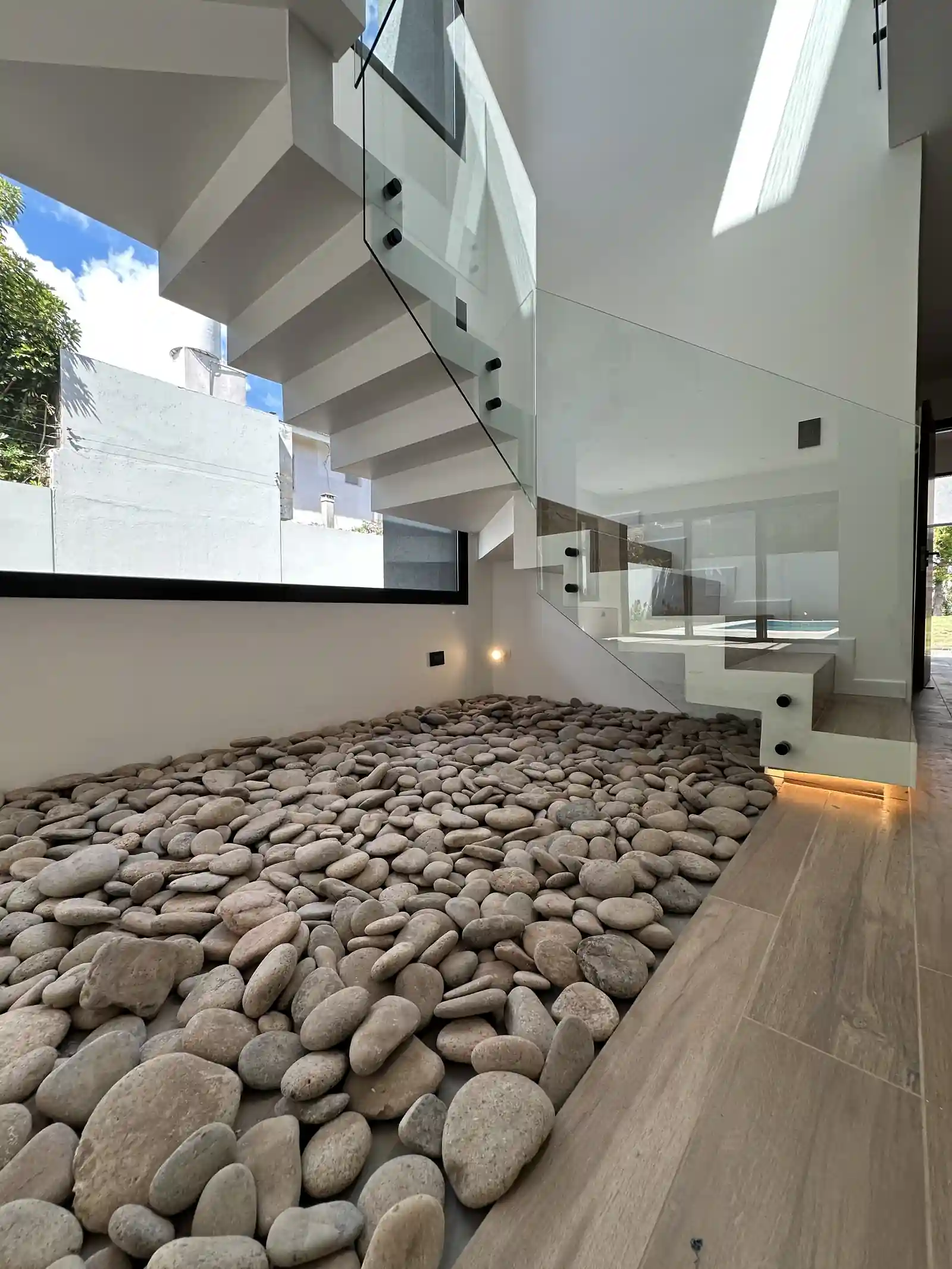 Escalera de madera y vidrio de Casa Adrogué sobre cantero interior de canto rodado, con doble altura